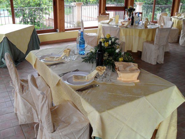 Tavoli del ristorante apparecchiati per la cena, con tovaglie giallo chiaro e centrotavola floreali, vicino a una finestra.