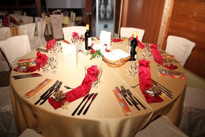Tavolo rotondo apparecchiato per una cena formale, con tovaglia dorata, tovaglioli rossi e bottiglie di vino.