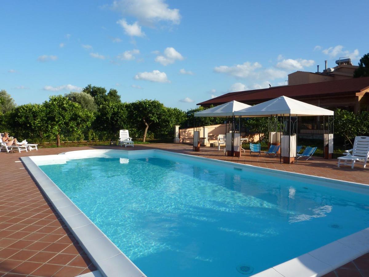 Piscina con acqua cristallina, lettini bianchi, ombrelloni e un edificio sotto un cielo azzurro.