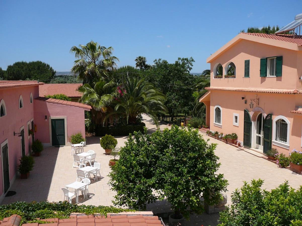 Cortile con edifici rosa, alberi, tavoli bianchi e cielo azzurro.