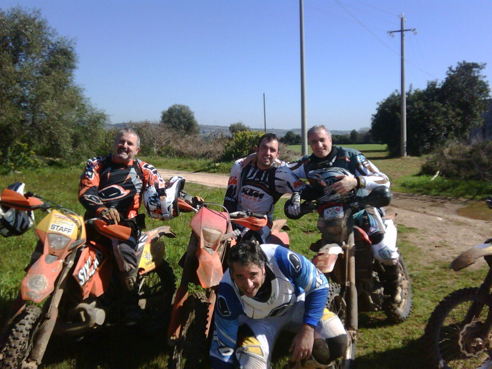 Quattro uomini in moto da cross, in posa all'aperto in una giornata di sole. Indossano caschi e abbigliamento da motociclista.