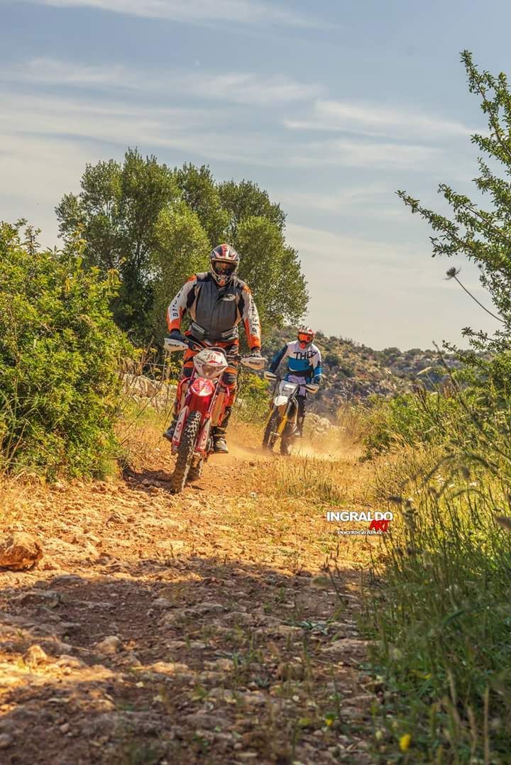 Due persone in sella a moto da cross su un sentiero roccioso, sollevando polvere. Illuminato dal sole, con alberi e cielo sullo sfondo.