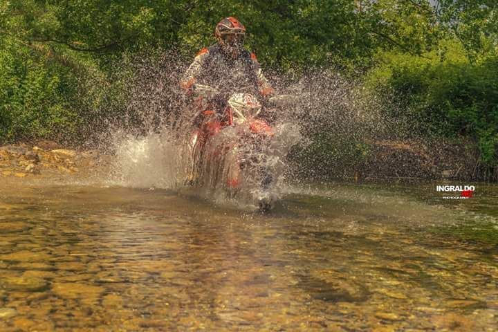 Un motociclista con la tuta rossa attraversa un ruscello poco profondo, creando grandi schizzi d'acqua.