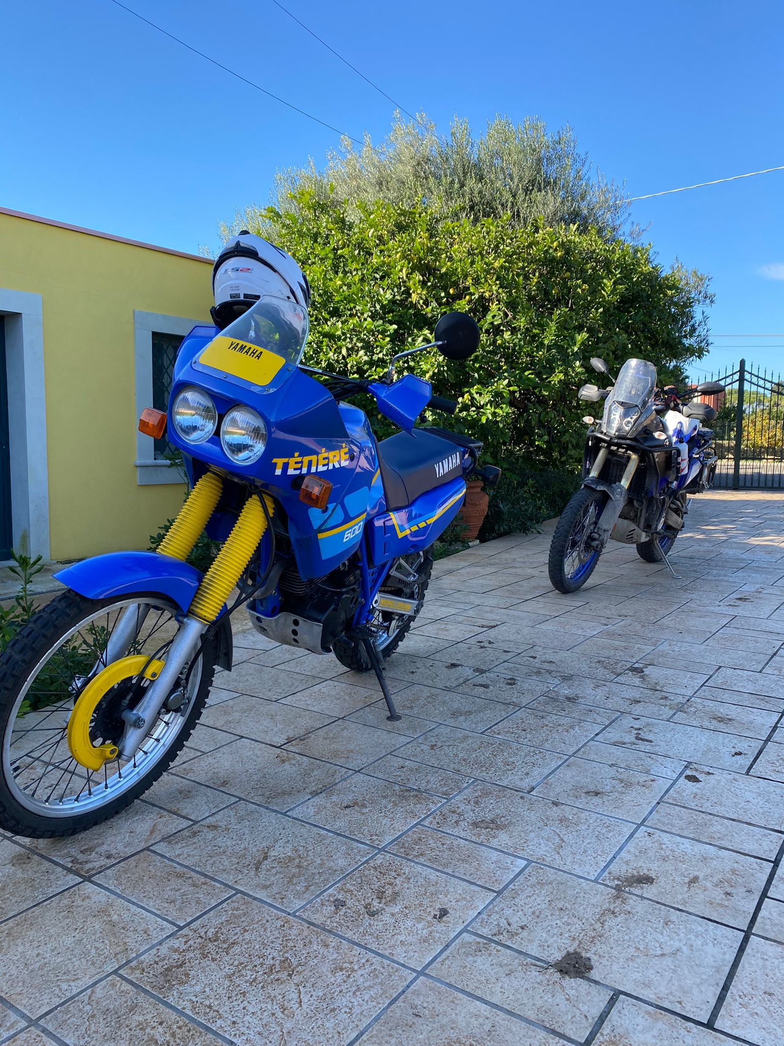Motocicletta KTM blu e gialla parcheggiata su una superficie di pietra, un'altra motocicletta sullo sfondo, giornata di sole.