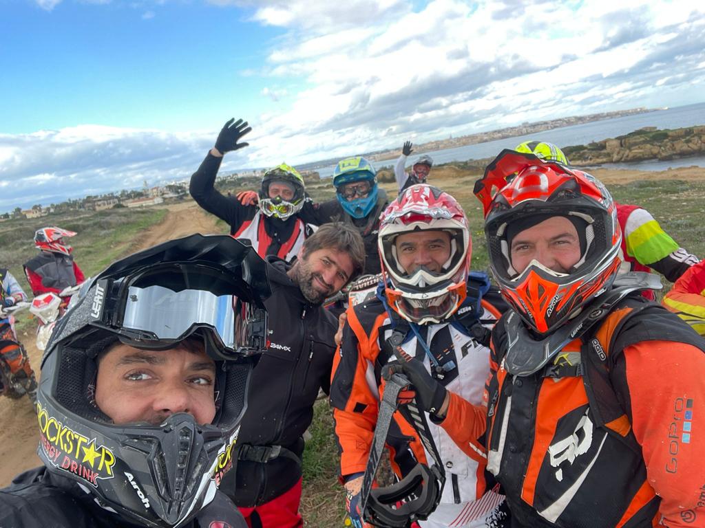 Un gruppo di motociclisti sorridenti scatta un selfie su una pista sterrata vicino alla costa.