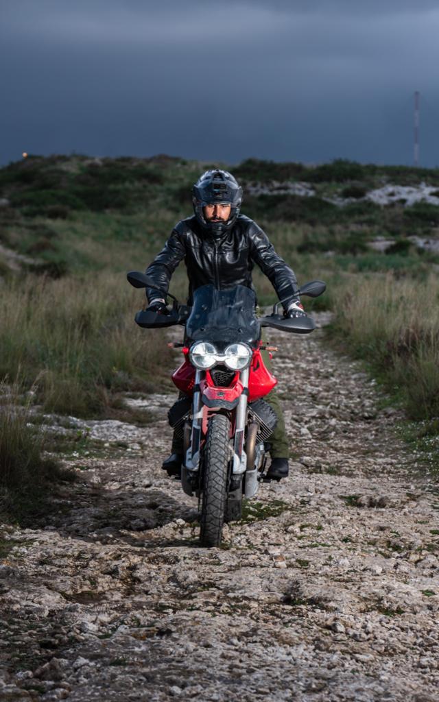 Un uomo su una motocicletta rossa percorre una strada sterrata, indossando casco e impermeabile. Sullo sfondo c'è un cielo scuro.