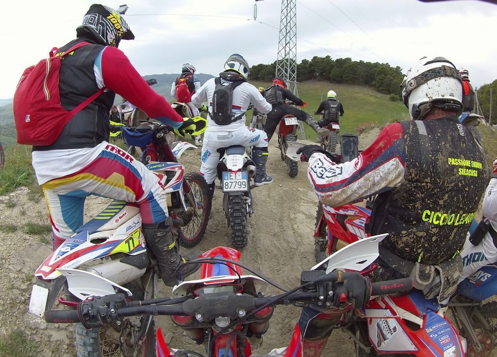 Motociclisti da cross su un sentiero fangoso, con indosso caschi e protezioni, pronti a pedalare in salita.