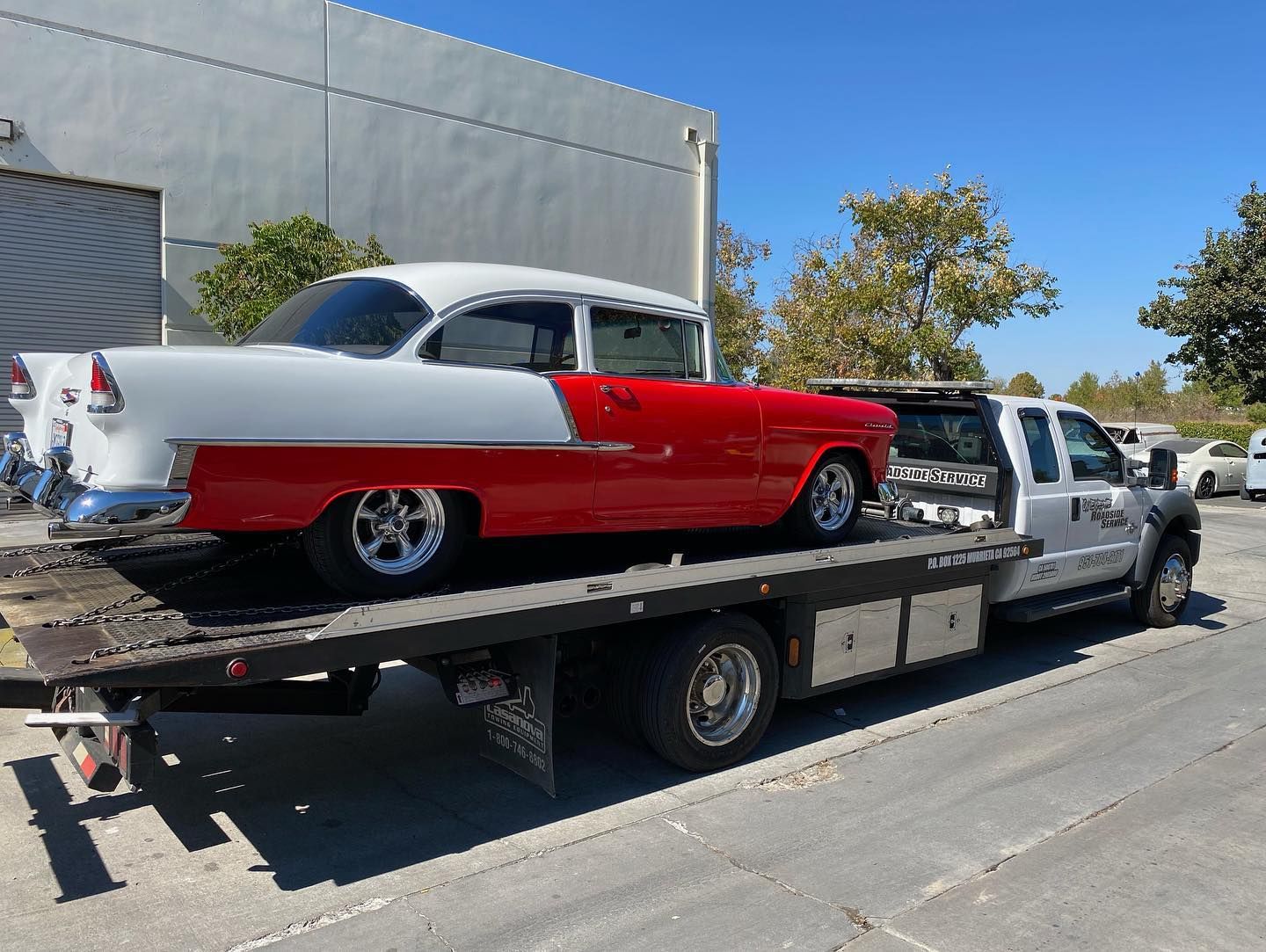 Classic red and white car on a flatbed tow truck, parked on a paved street, sunny day.