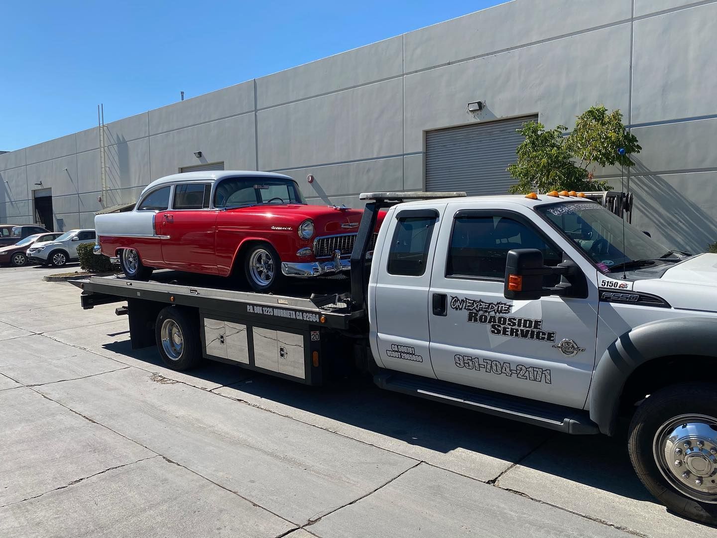 Red and white classic car being transported on a tow truck. Outdoors in front of a building on a sunny day.