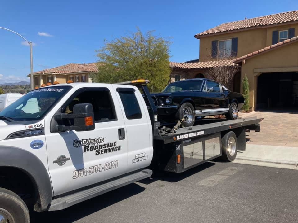 Black classic car on a tow truck in front of a house on a sunny day.