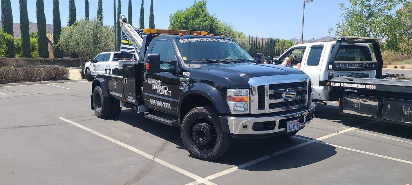 A black tow truck parked in a lot next to a white flatbed tow truck on a sunny day.