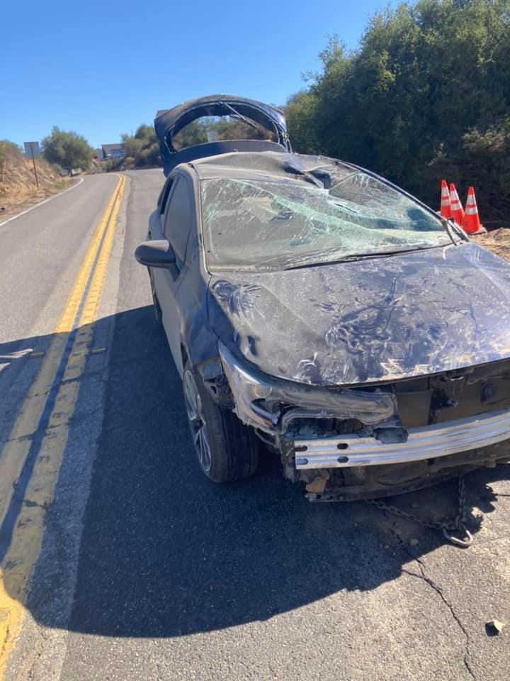Damaged blue car on a road, windshield and roof severely damaged, trunk open.