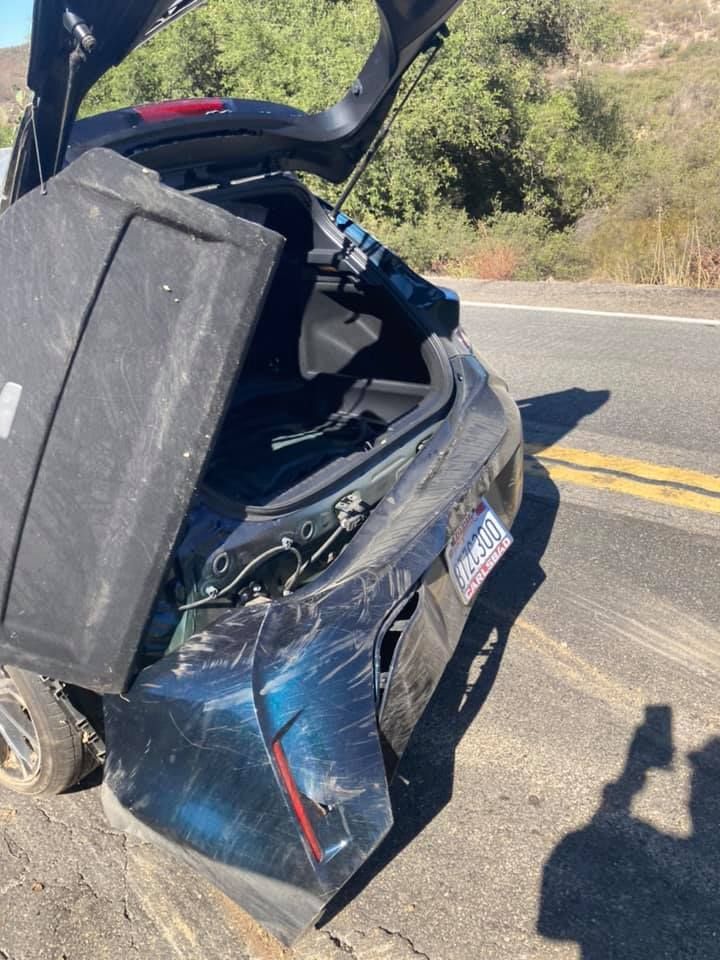 Damaged blue car with trunk open on side of a road after an accident.