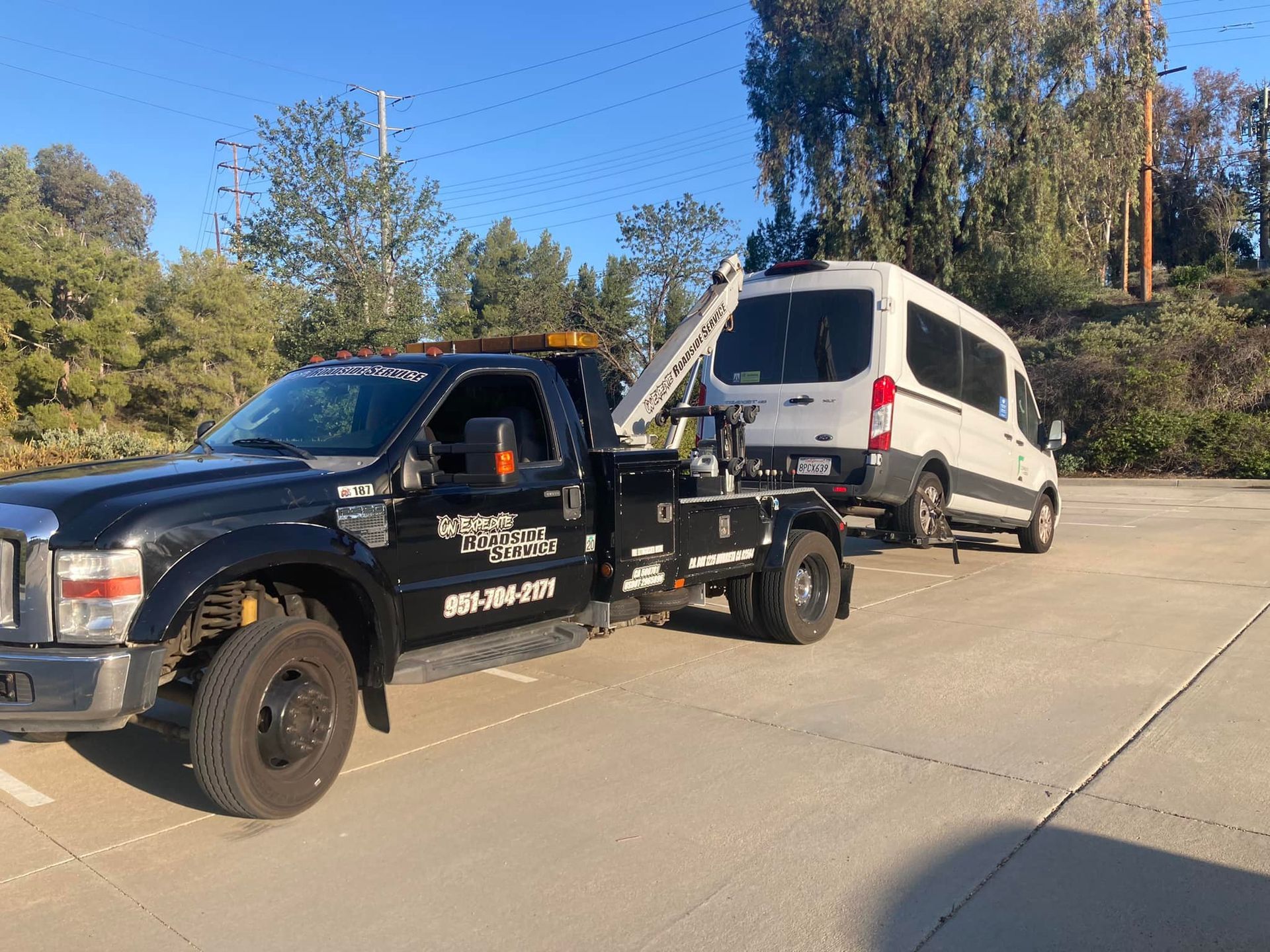 Black tow truck towing a white van on a paved lot, trees in the background, blue sky.