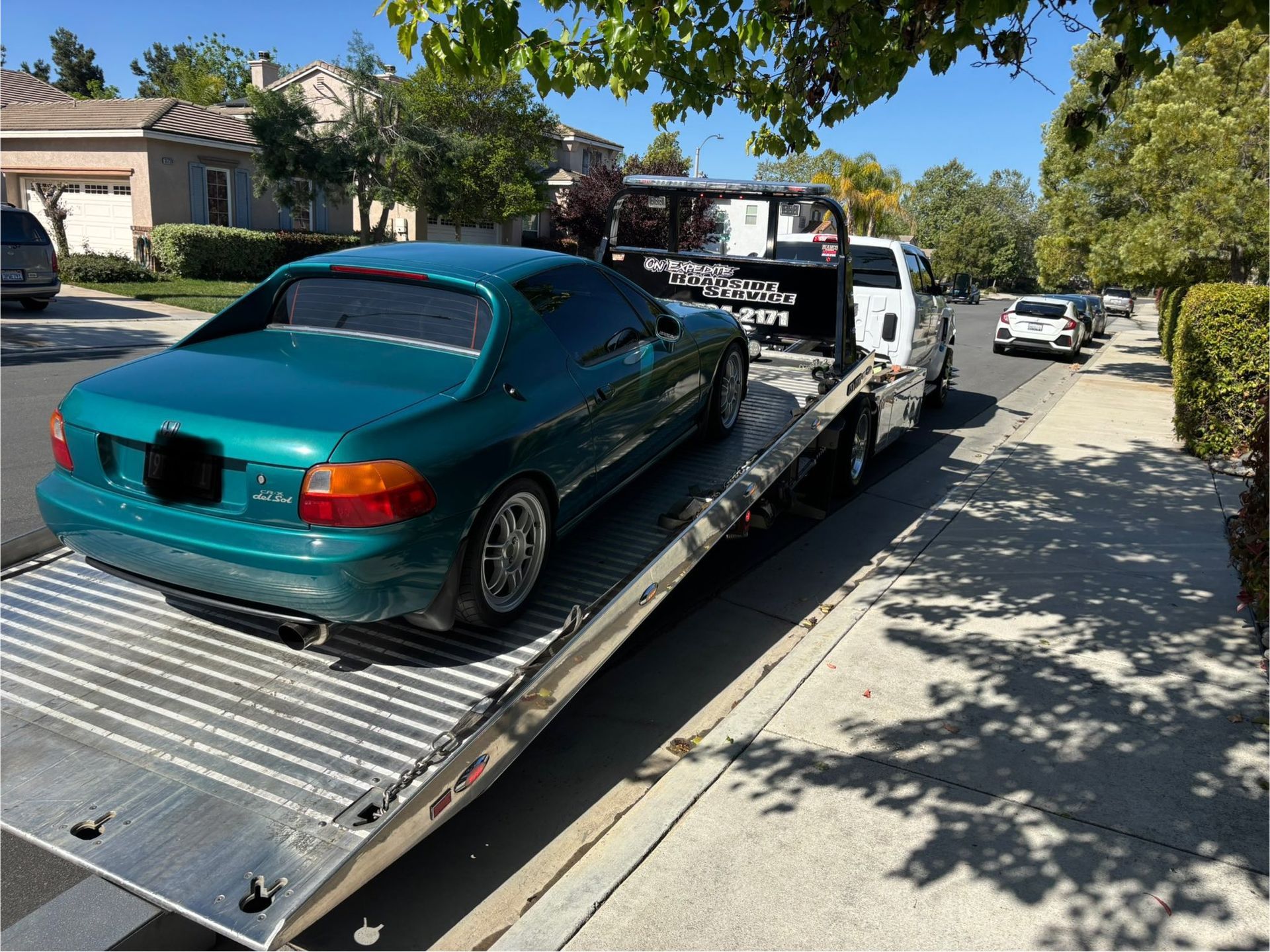 A teal coupe on a flatbed tow truck on a sunny residential street.