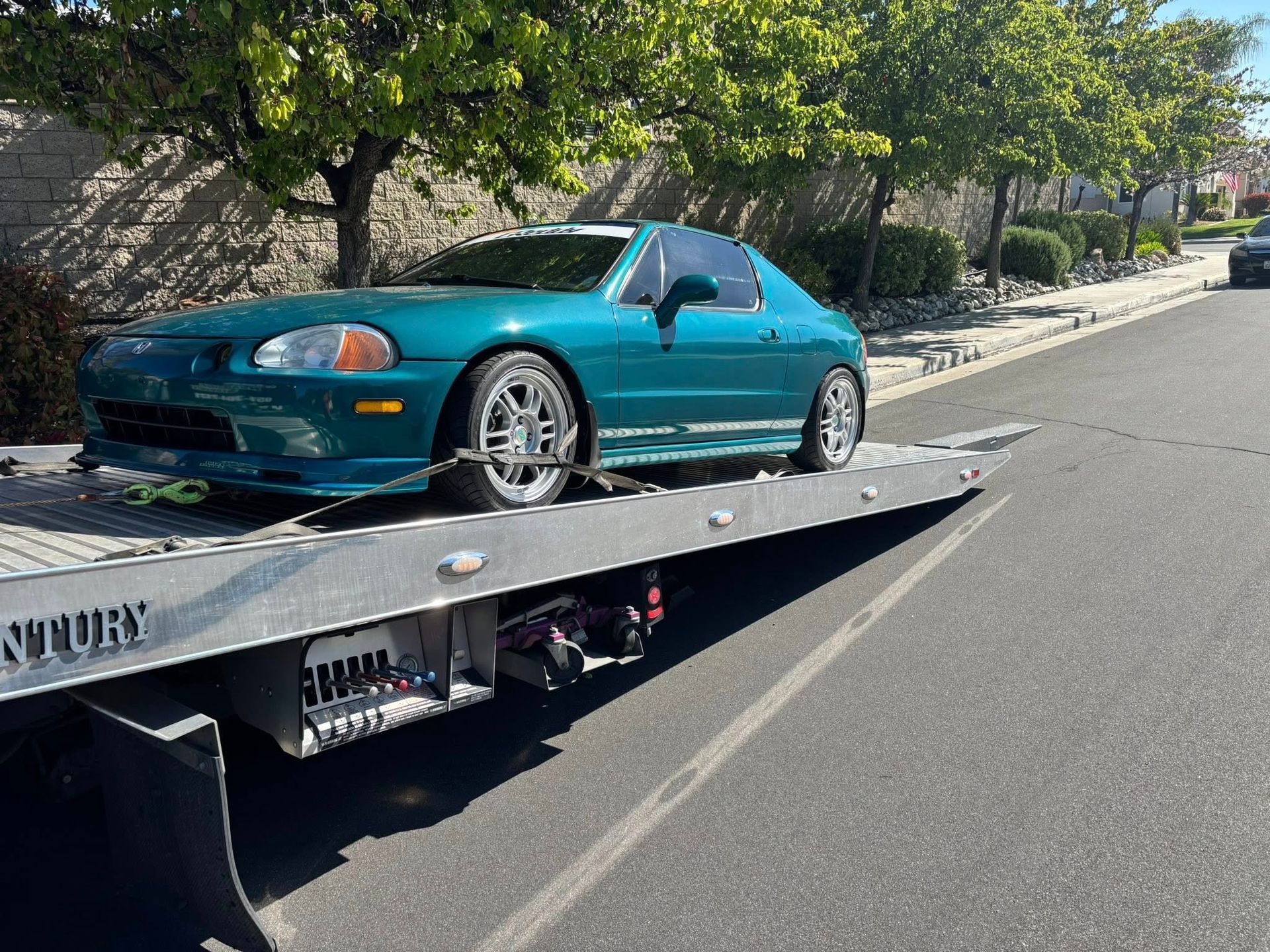 Green Honda Del Sol on a flatbed tow truck, parked on a residential street.