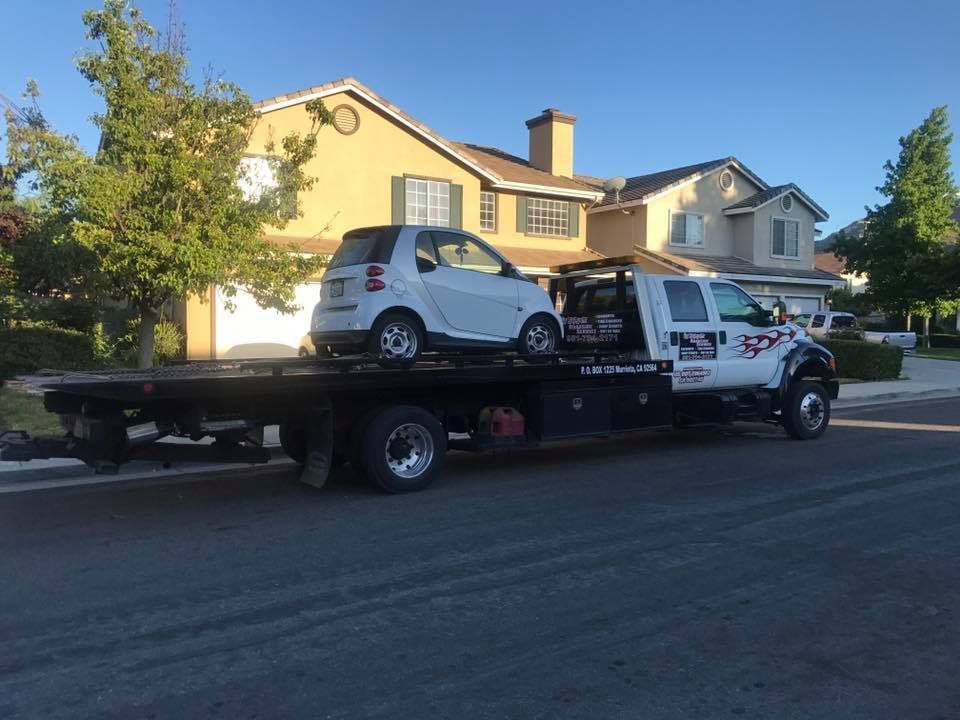 A white Smart car loaded on a flatbed tow truck, parked on a residential street.