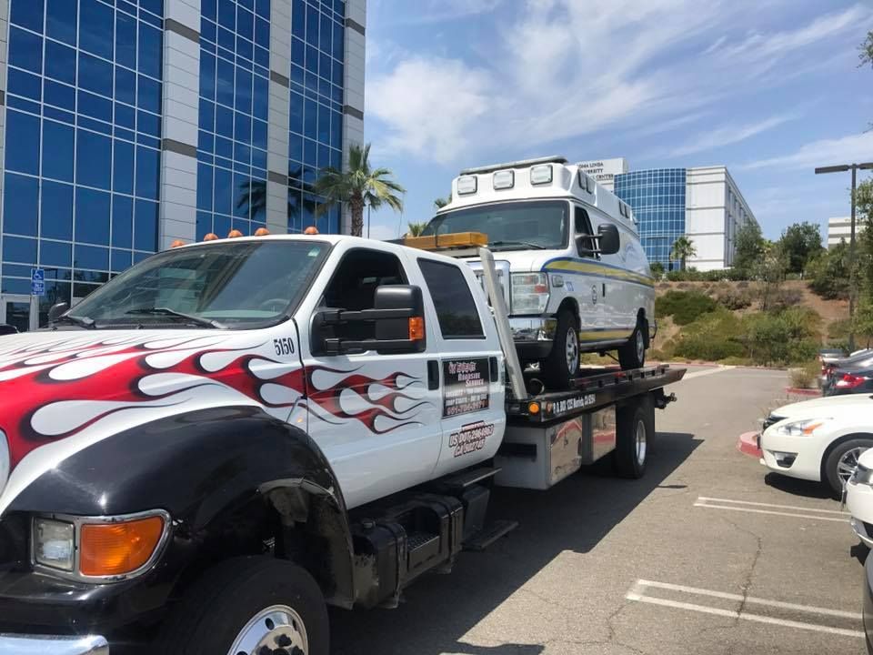 Tow truck hauling an ambulance, parked in front of a modern building under a blue sky.