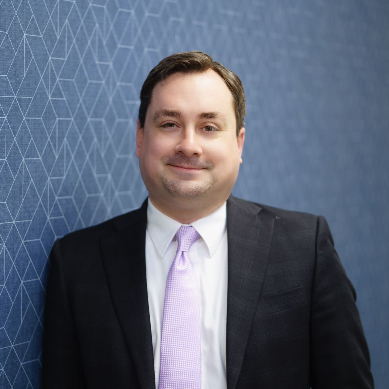 Smiling man (Ben Yelverton) in a dark suit and light purple tie against a blue patterned backdrop