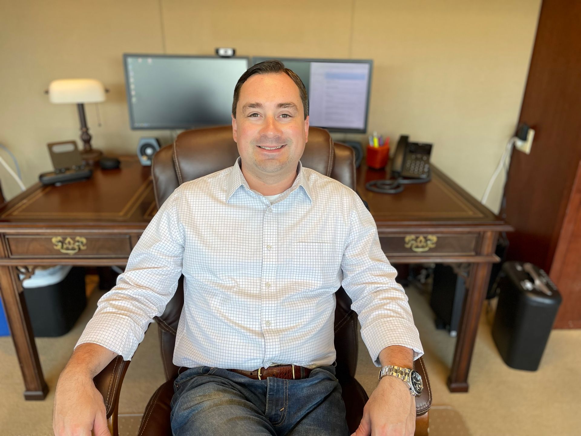 Man seated in an office chair at a desk, smiling, with a computer monitor and lamp behind him.