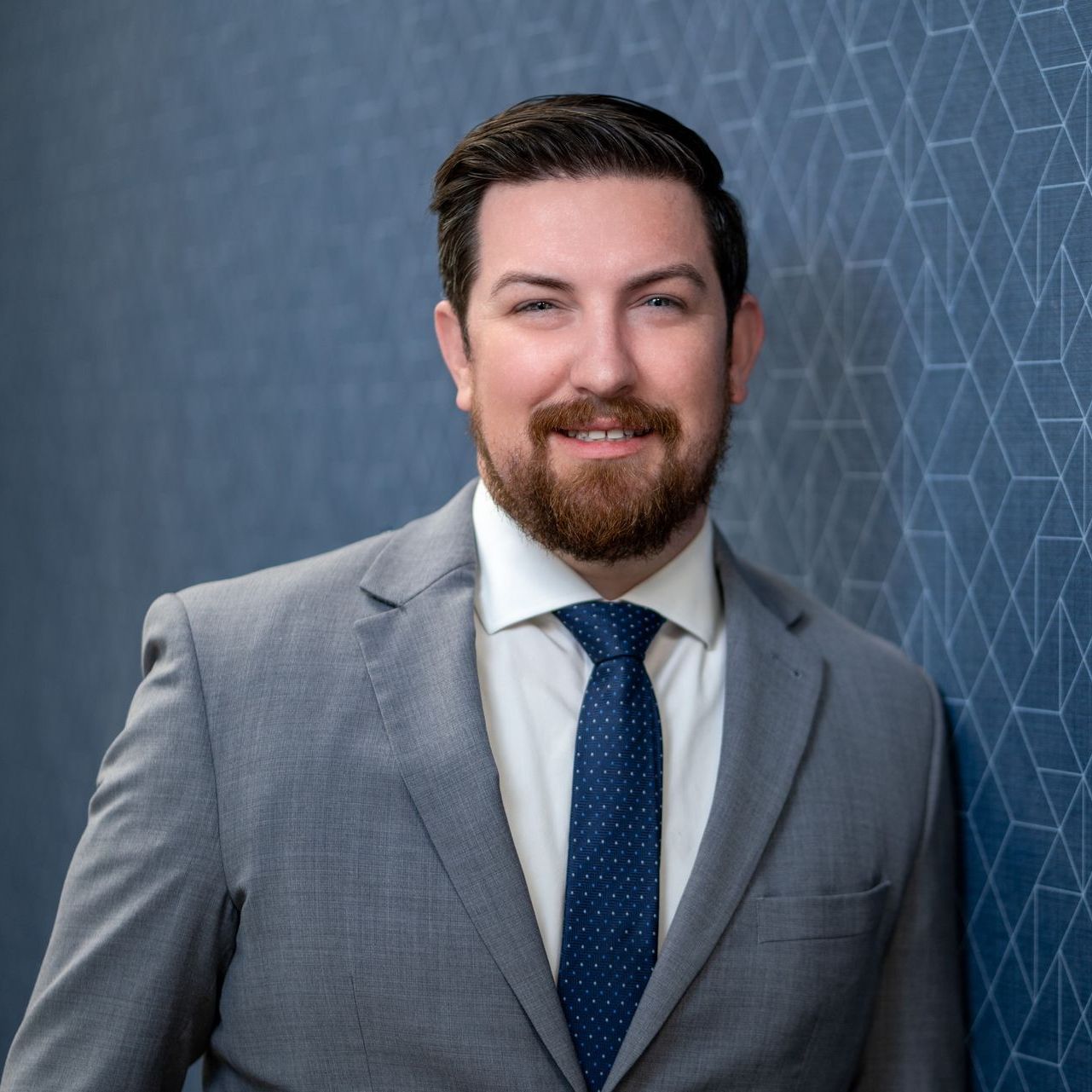 Smiling man in a gray suit and blue tie against a blue geometric wall