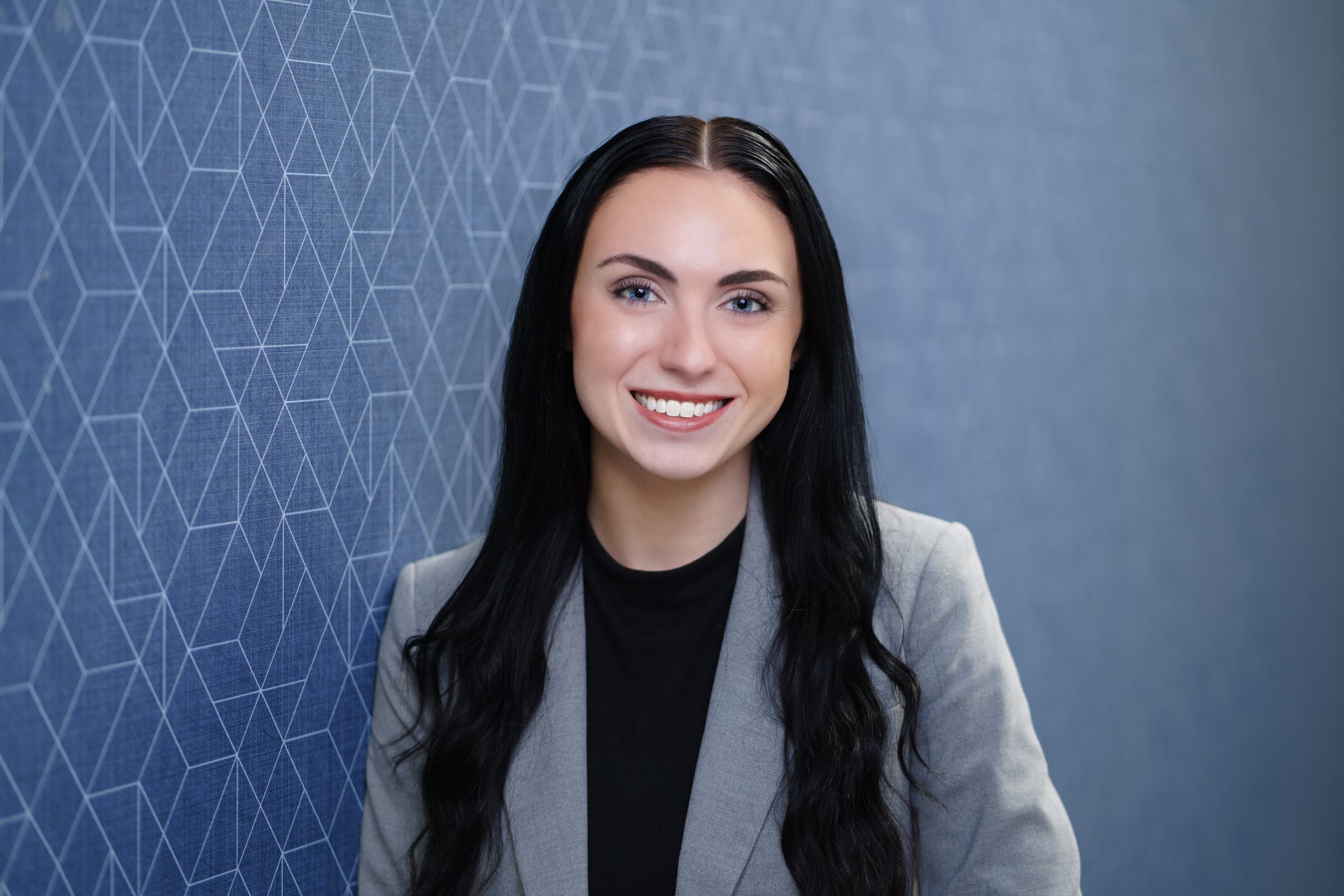 Smiling woman (Olivia Blackford) in a gray blazer against a blue patterned wall