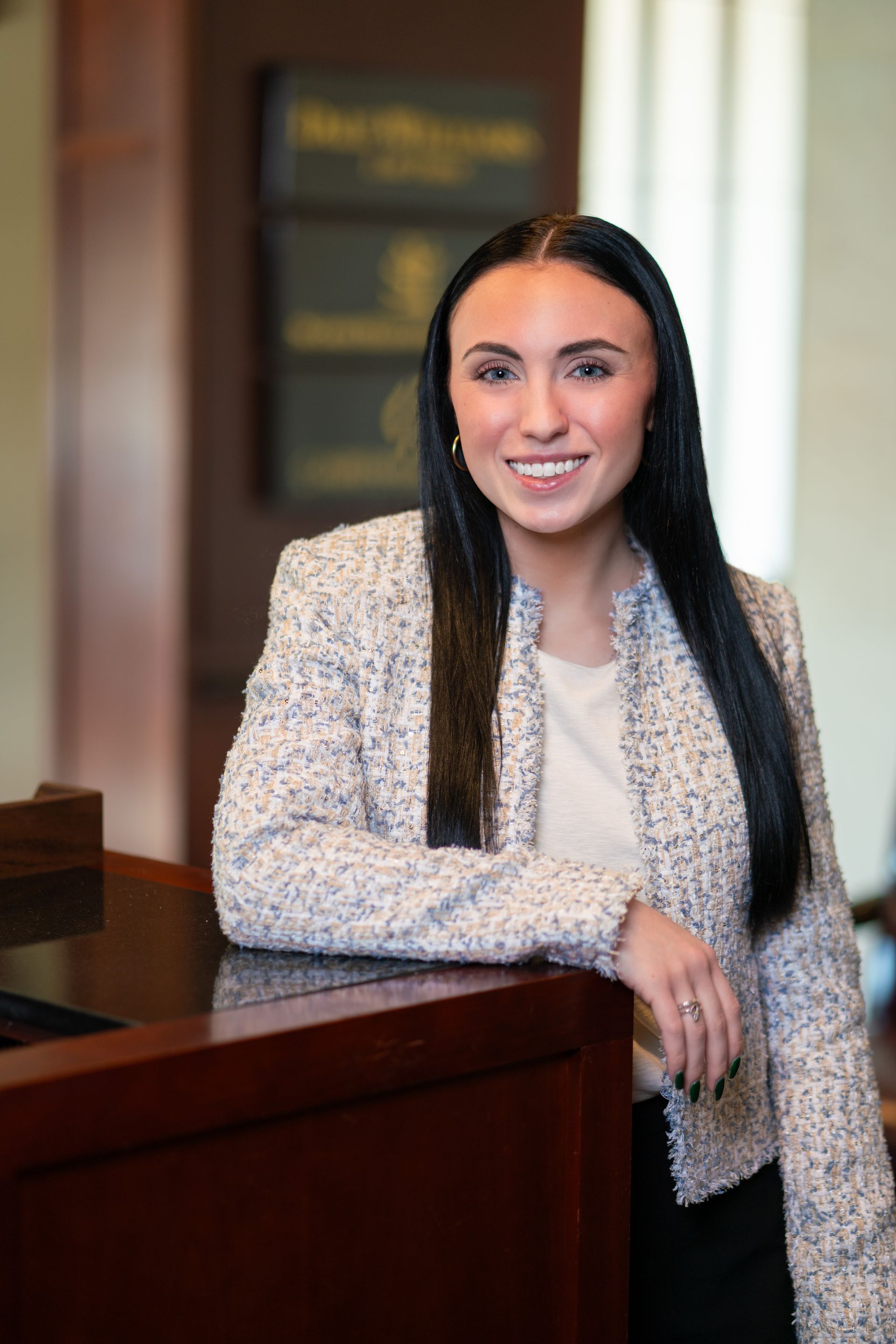 Woman smiling beside a wooden desk in a bright office, wearing a light tweed blazer.