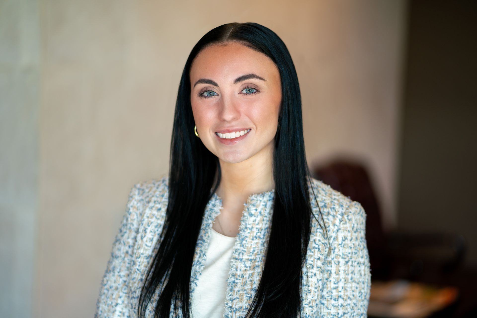 Portrait of a smiling woman with long dark hair in a light textured jacket, standing indoors.