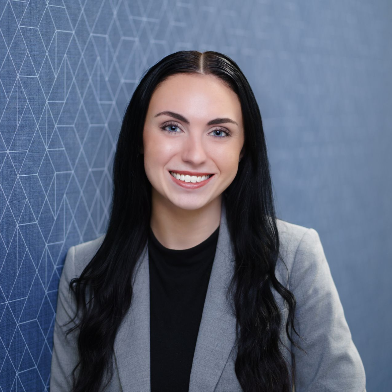 Smiling women (Olivia Blackford) with long dark hair in a gray blazer against a blue patterned background