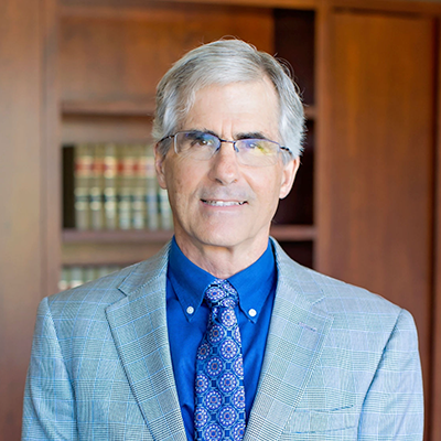 Headshot of a man in a light gray suit and blue shirt, standing in an office setting.