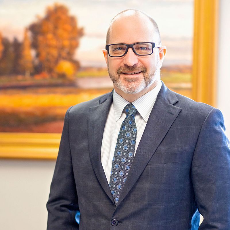 Man in a dark suit and blue tie smiling in an office, with a framed landscape painting behind him.