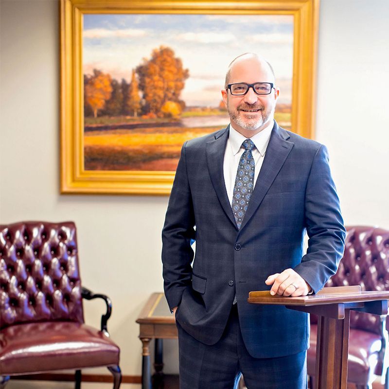 Man in a dark suit standing beside a wooden desk in an office, with a framed landscape painting behind him.