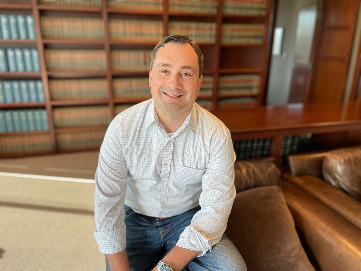 Smiling man in a patterned shirt sitting on a brown couch in a wood-paneled library