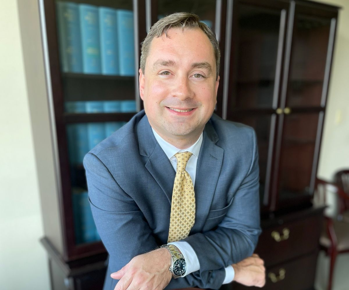 Smiling man in a blue suit with a yellow tie, leaning on a desk in an office.