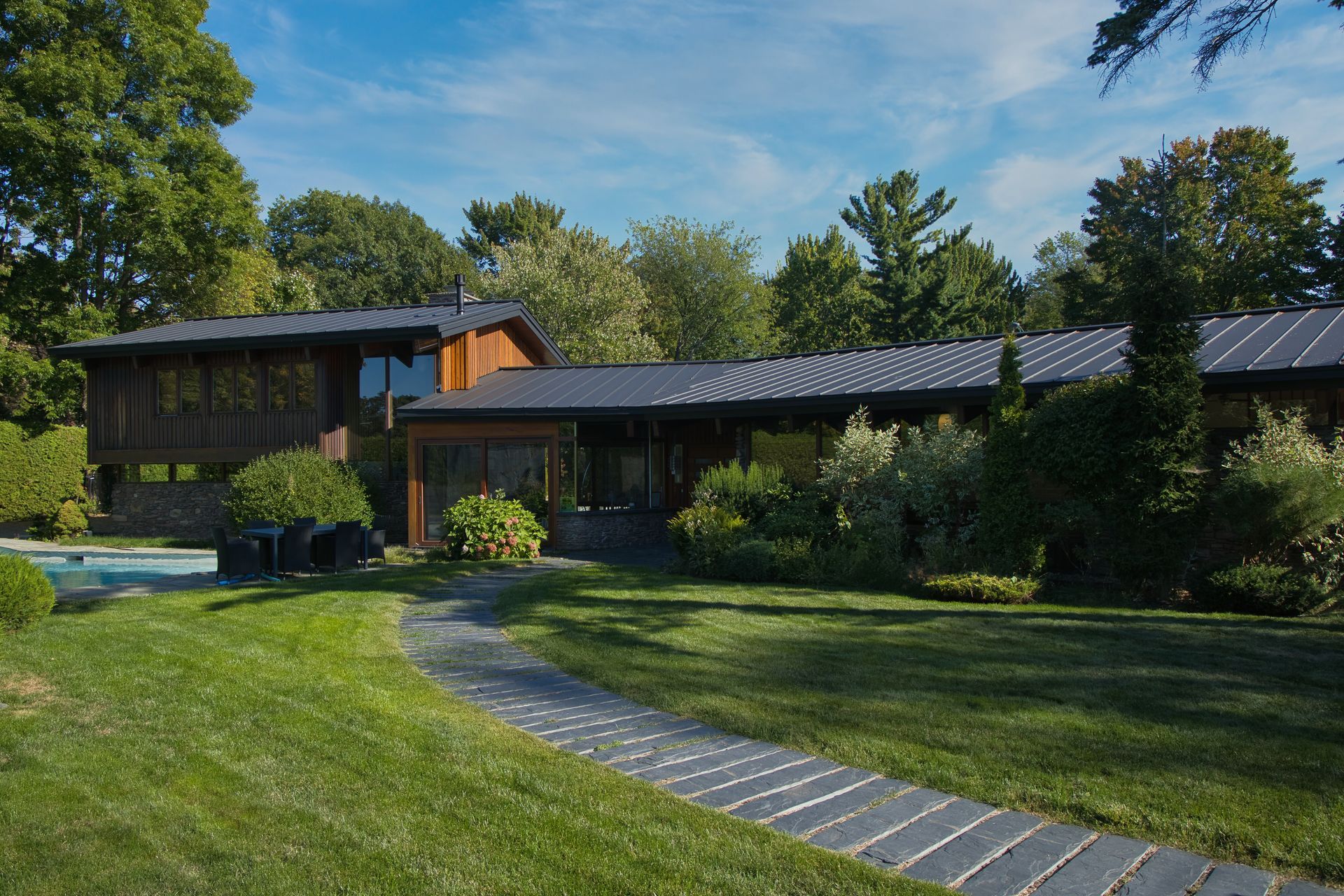 House with wooden siding and dark roof, set on green lawn with stone path. Trees in background.