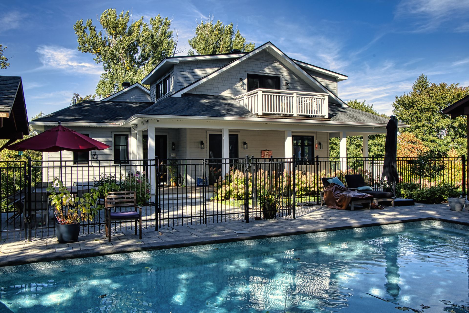 House with pool, covered porch, and balcony, behind a black fence. Sunny day.