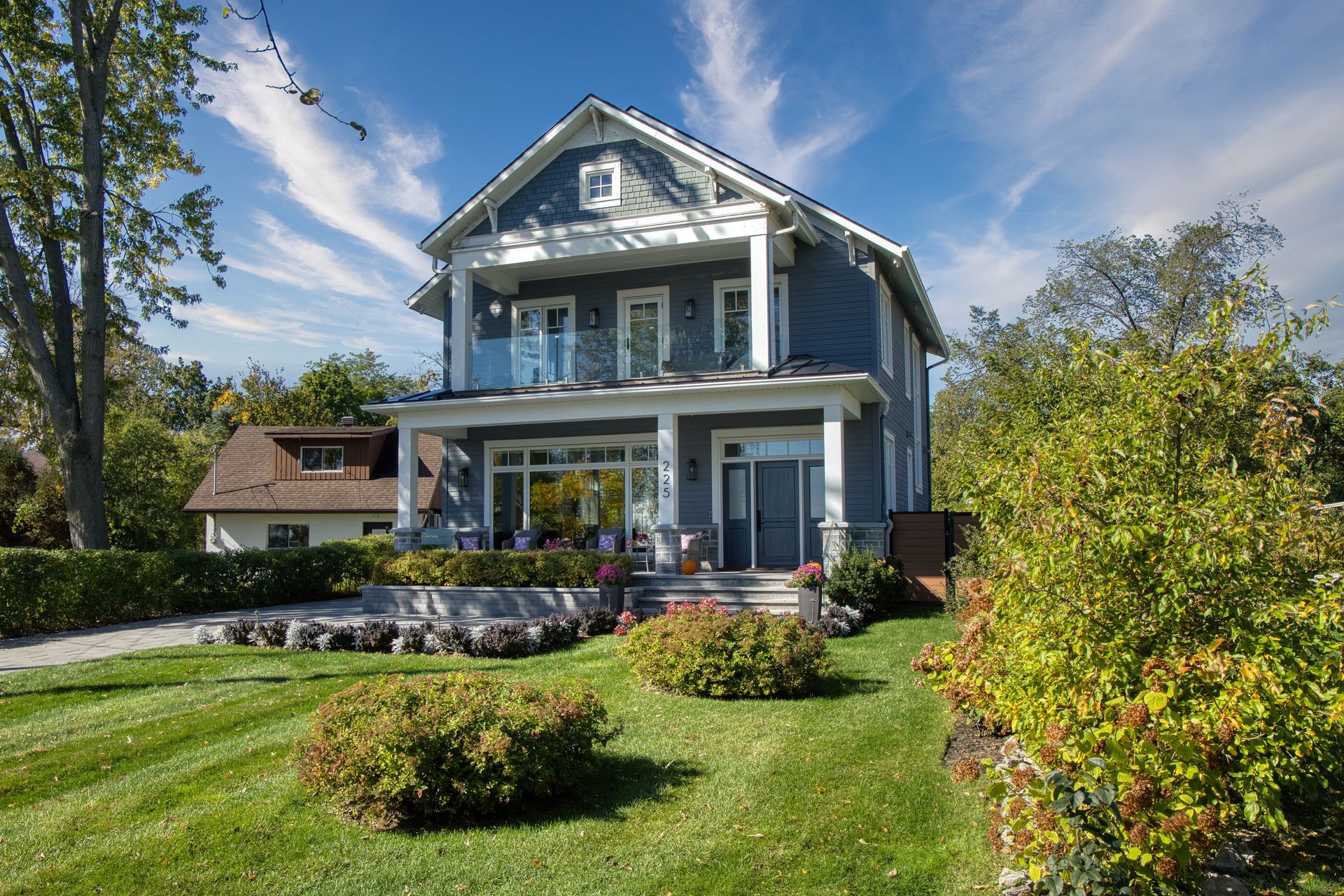 Two-story blue house with porch and balcony, set on a green lawn with shrubs, under a blue sky.