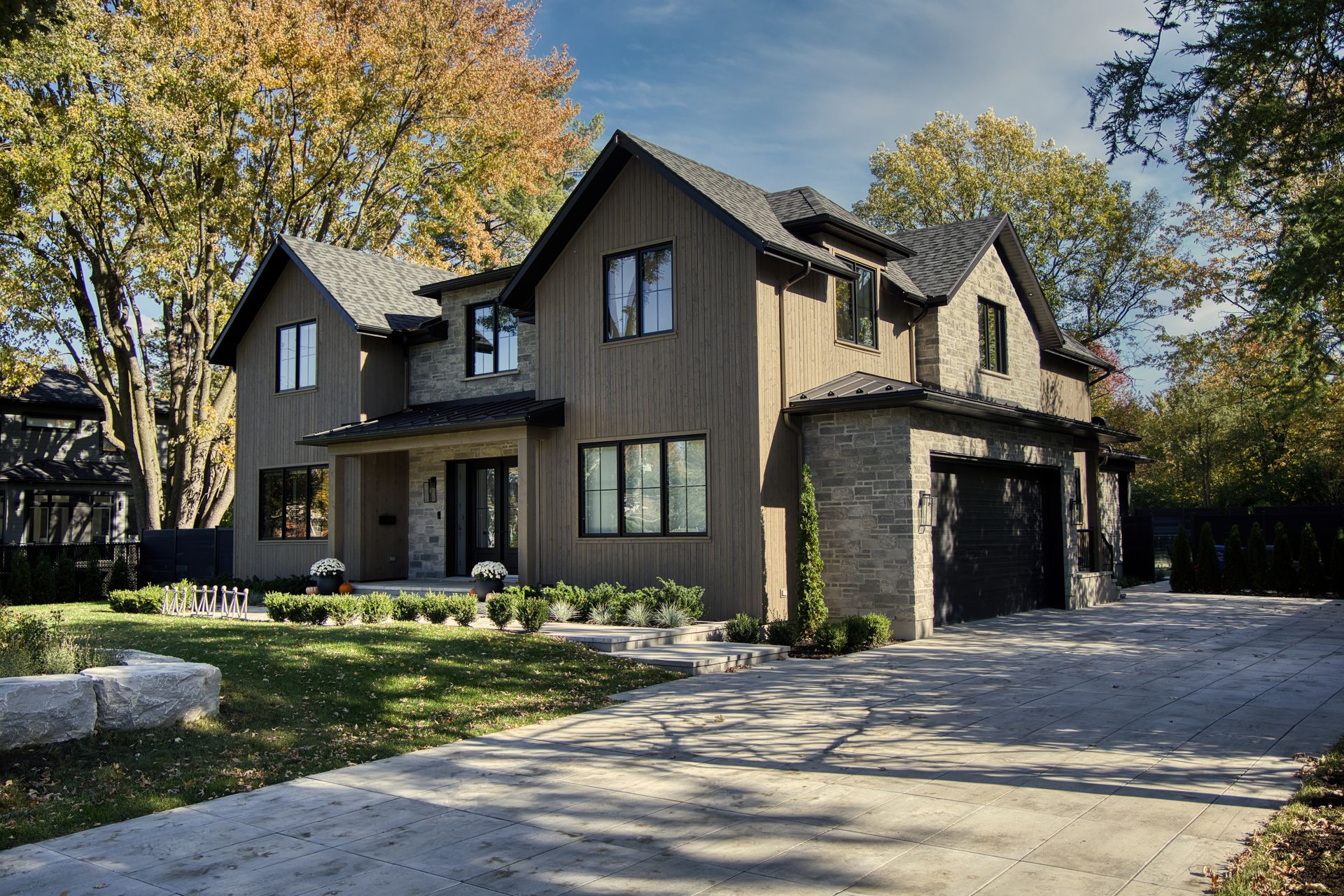 Two-story house with stone and wood siding, black windows, and a driveway, surrounded by trees.