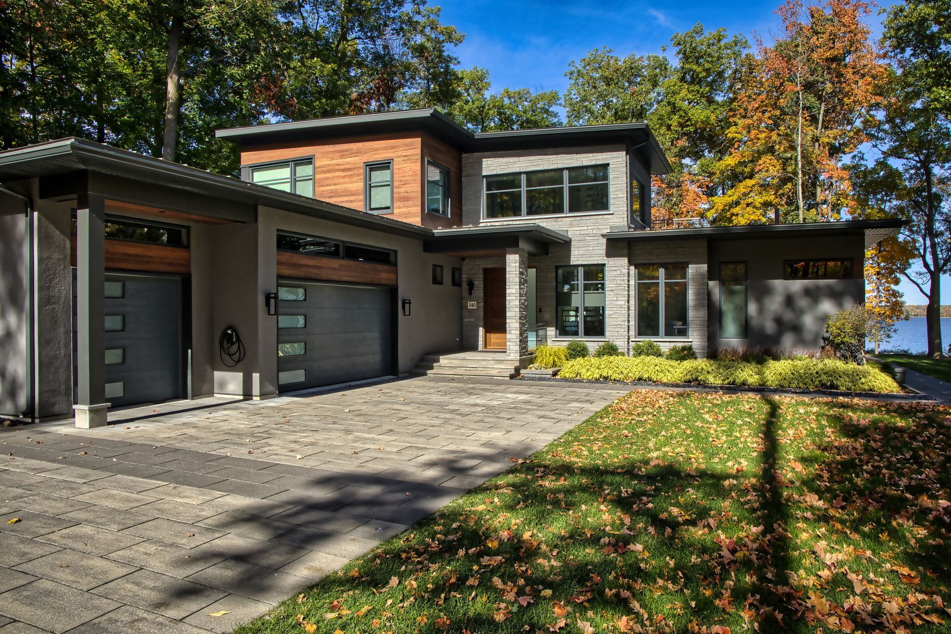 Modern two-story house with gray facade, wood accents, and a stone driveway. Trees and water in the background.