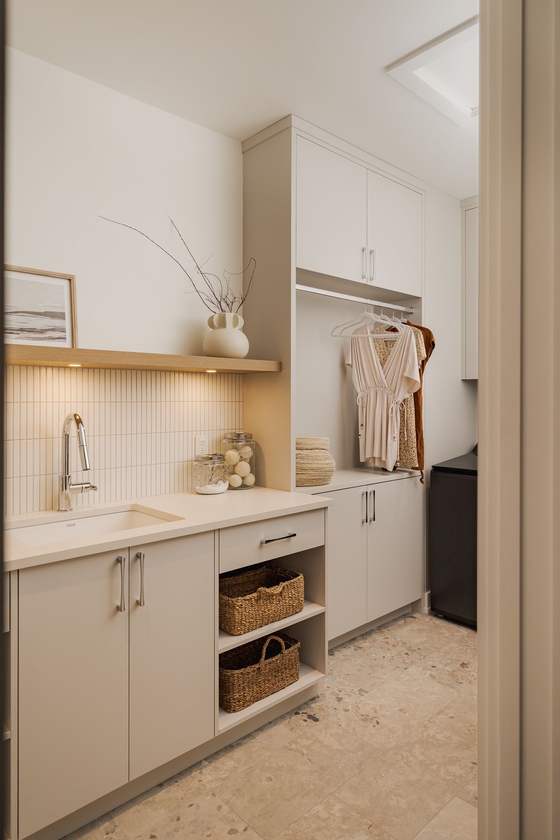 Laundry room with gray cabinets, sink, and clothes hanging. Light beige tile and flooring.