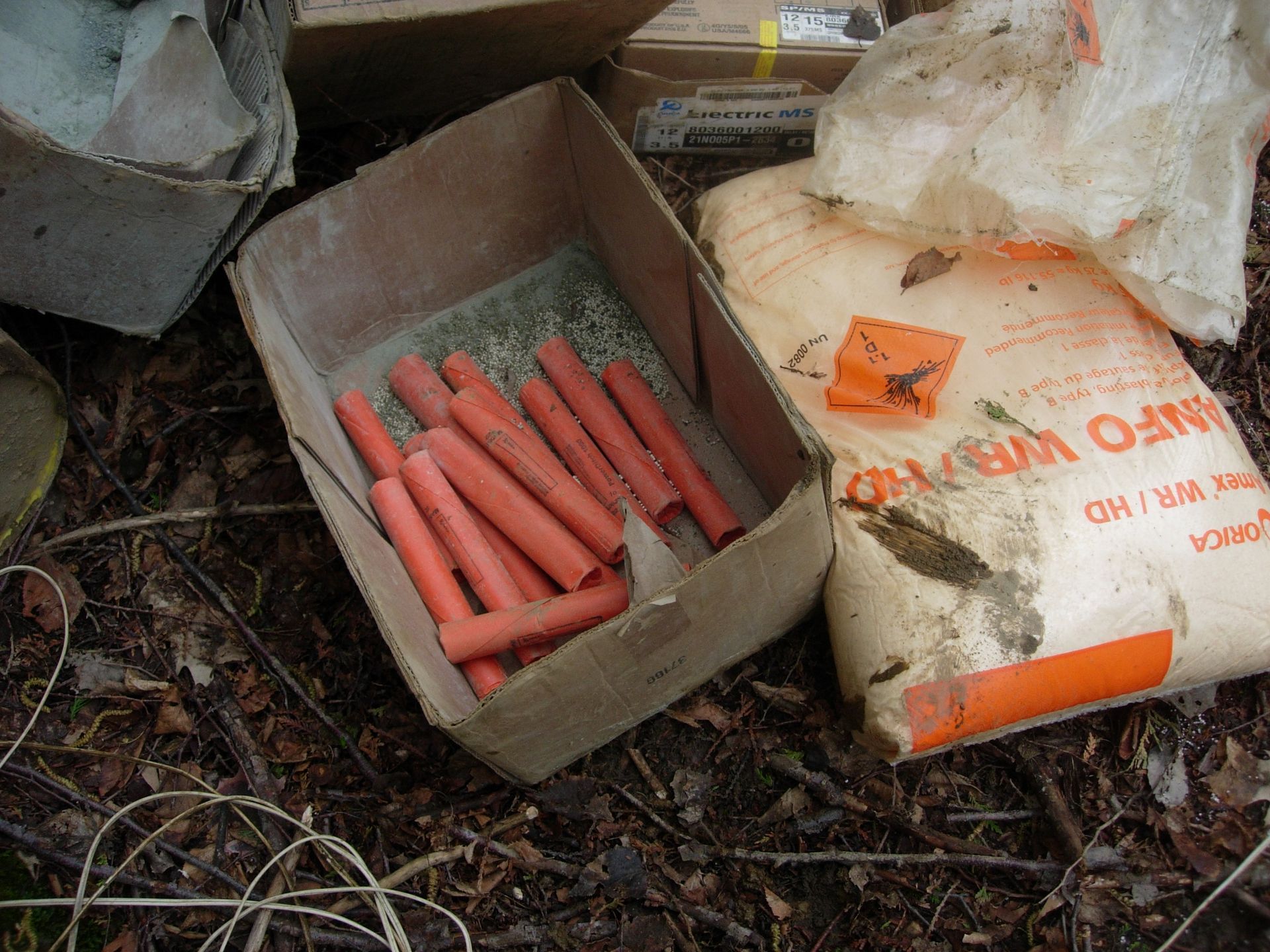 Cardboard box with red sticks, and a white bag with orange label, on forest floor.