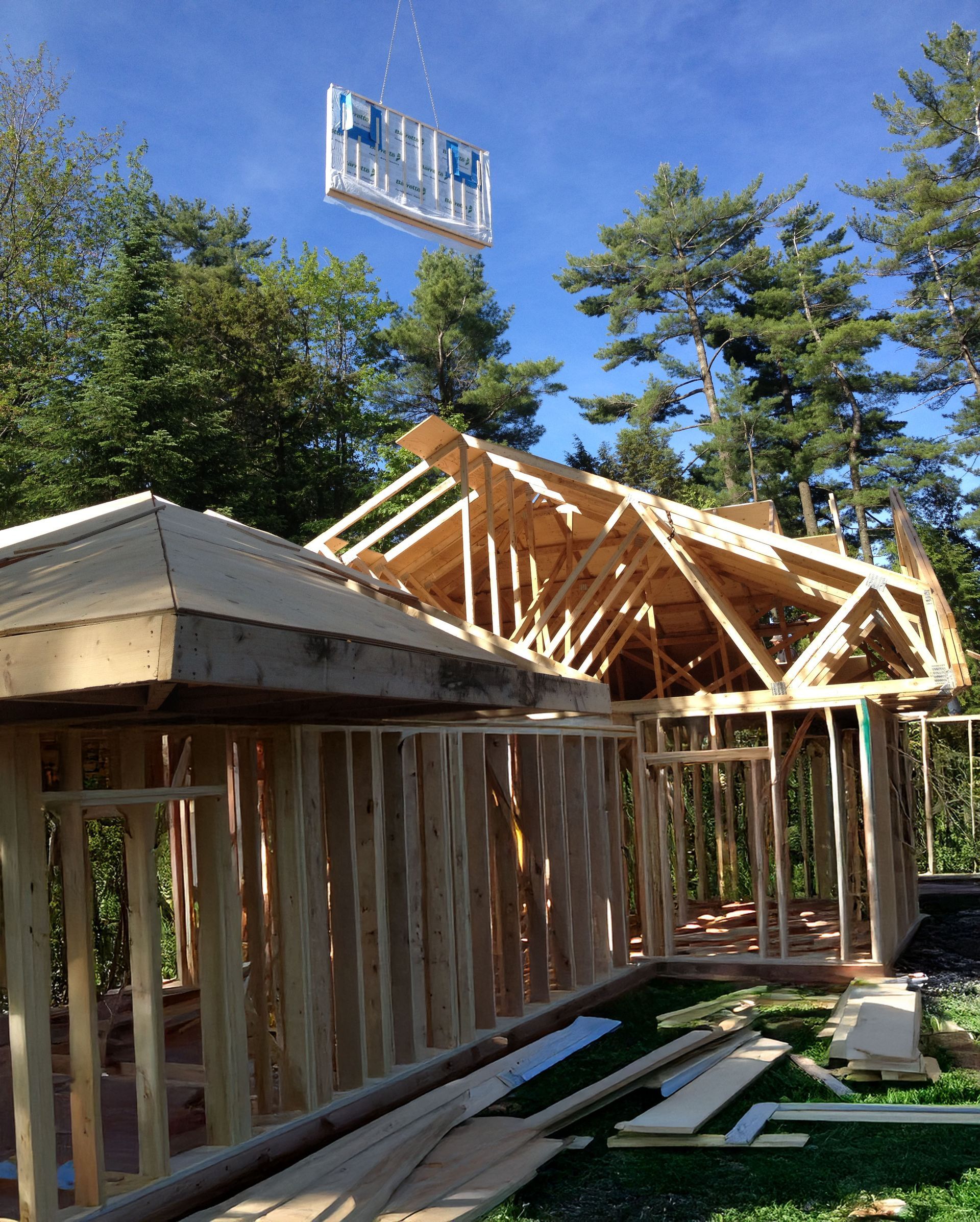 Construction site with wooden framing and a panel being lifted by a crane. Trees in the background.