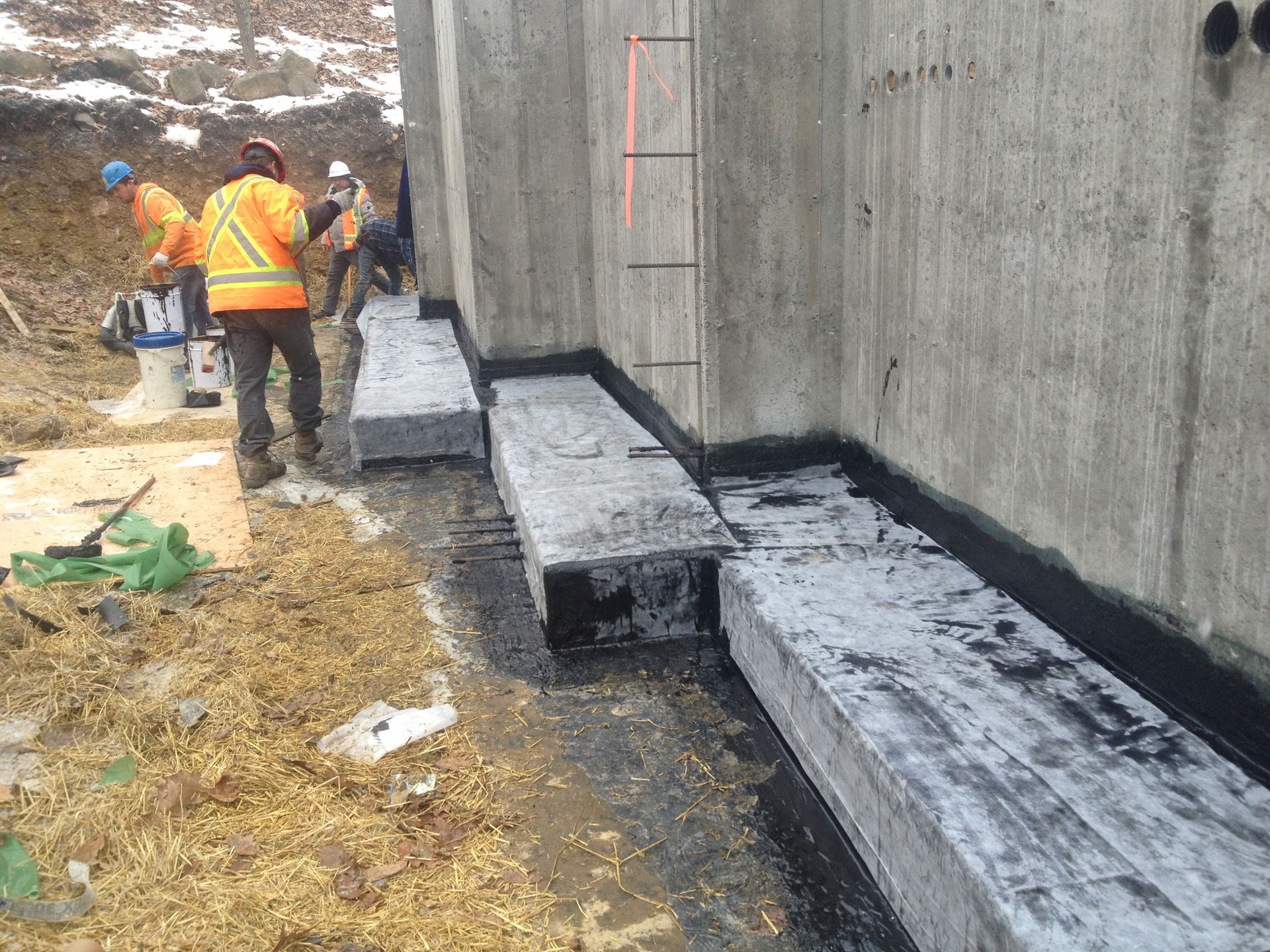 Construction workers applying sealant to concrete foundation walls outdoors.