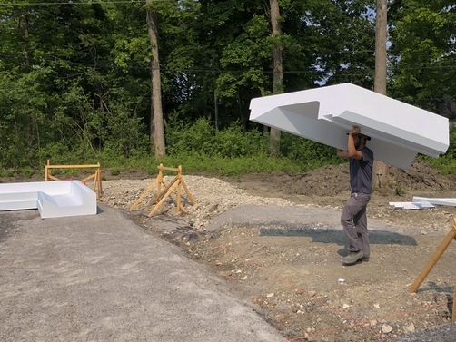 Man carries a large, white foam structure on his shoulders at a construction site near trees.