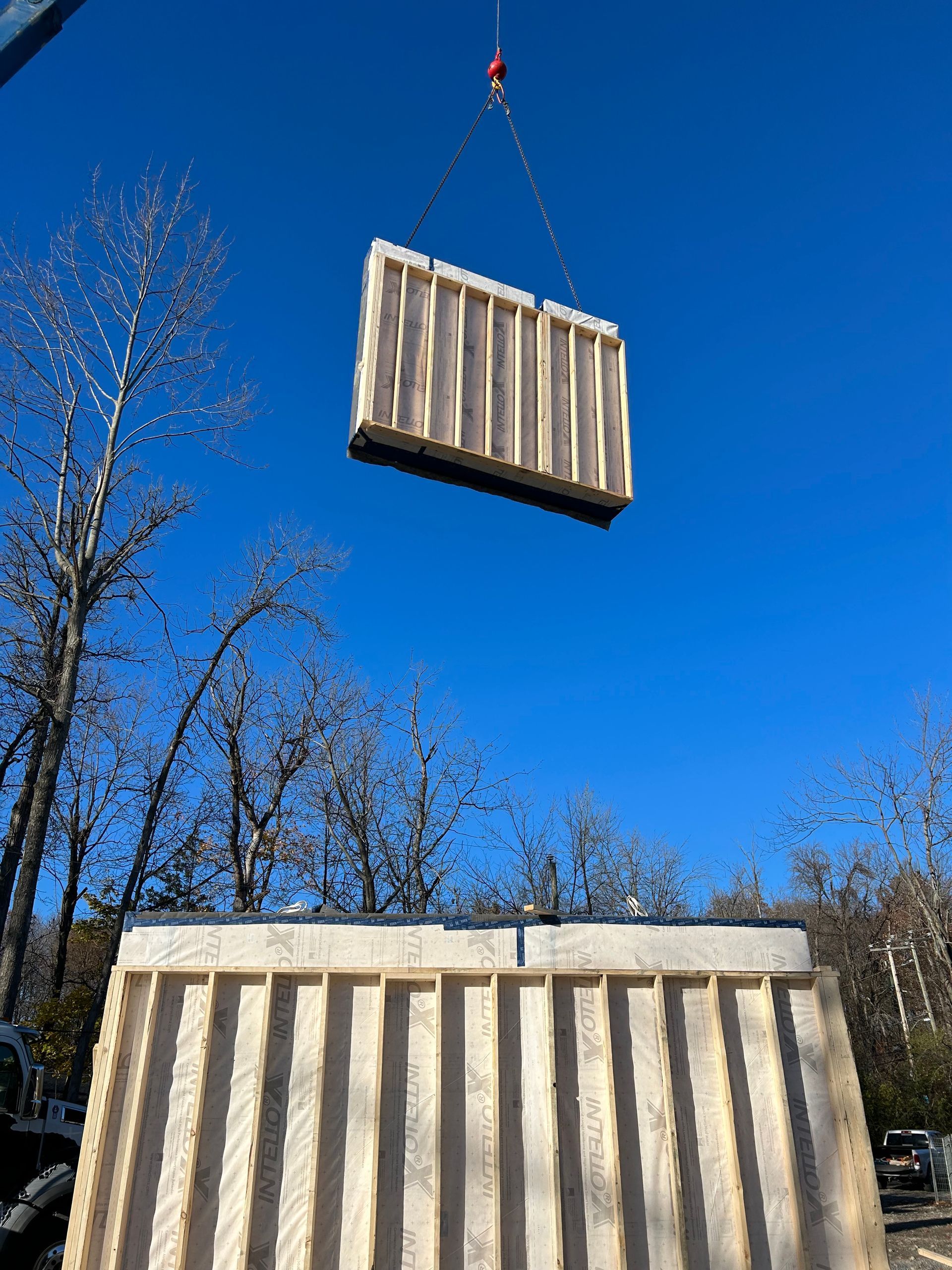 Crane lifting a wooden wall panel above another panel, against a bright blue sky.