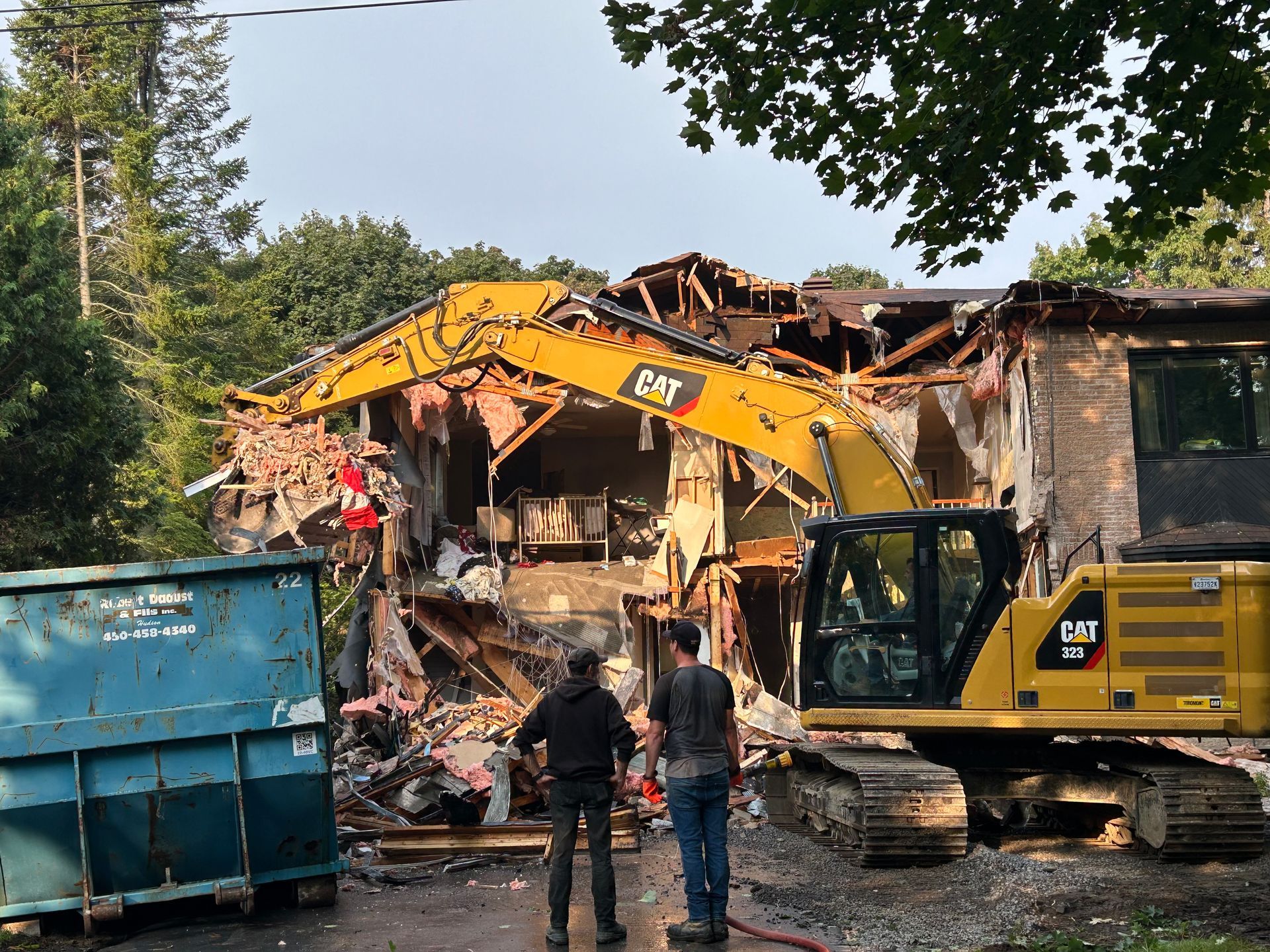 Demolition of a house; a yellow excavator tears through the structure; two men watch. A blue dumpster is in the foreground.