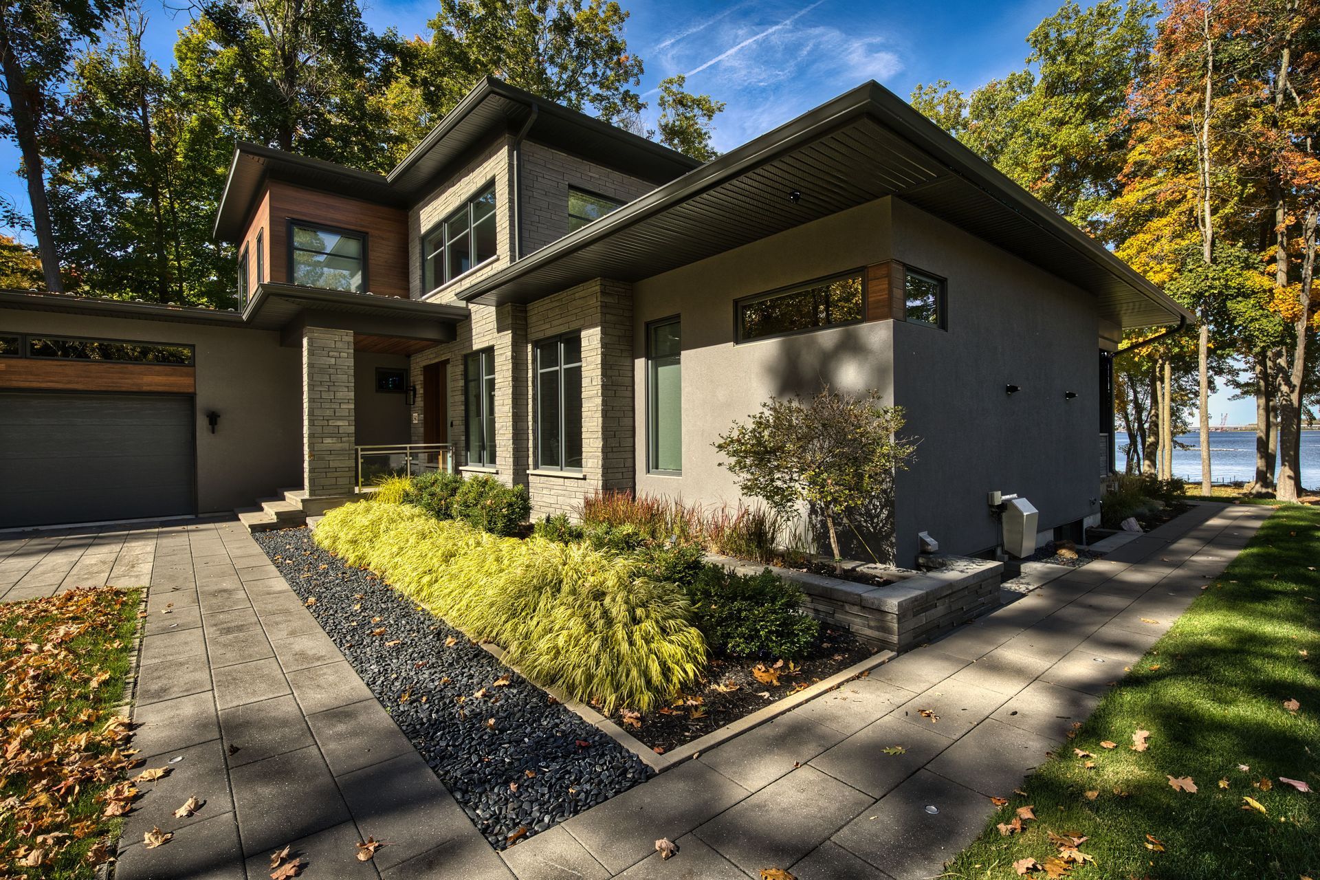 Modern two-story house with stone and gray exterior, lush landscaping, and a waterfront view.