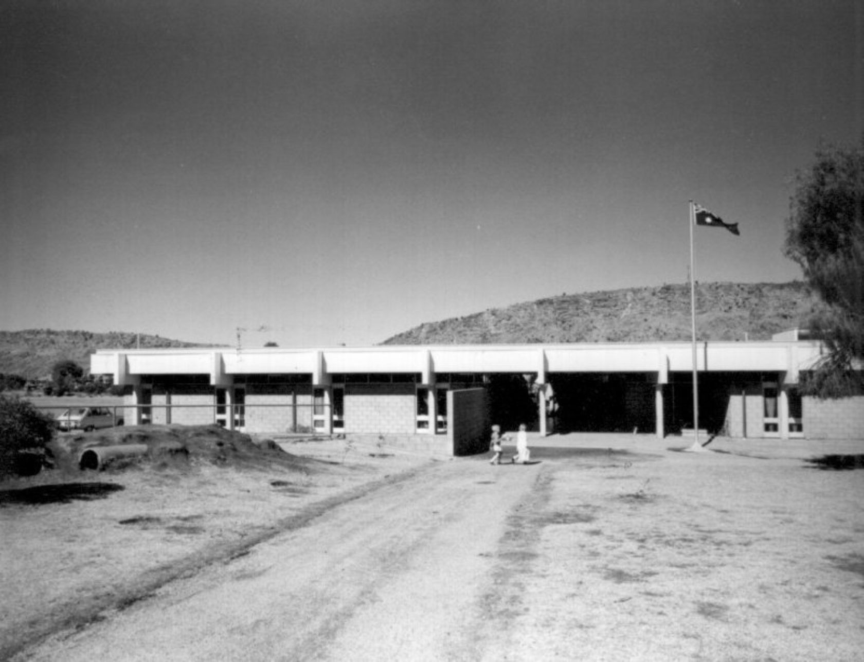 A black and white photo of a low, rectangular building with a flag in front, in a desert landscape.
