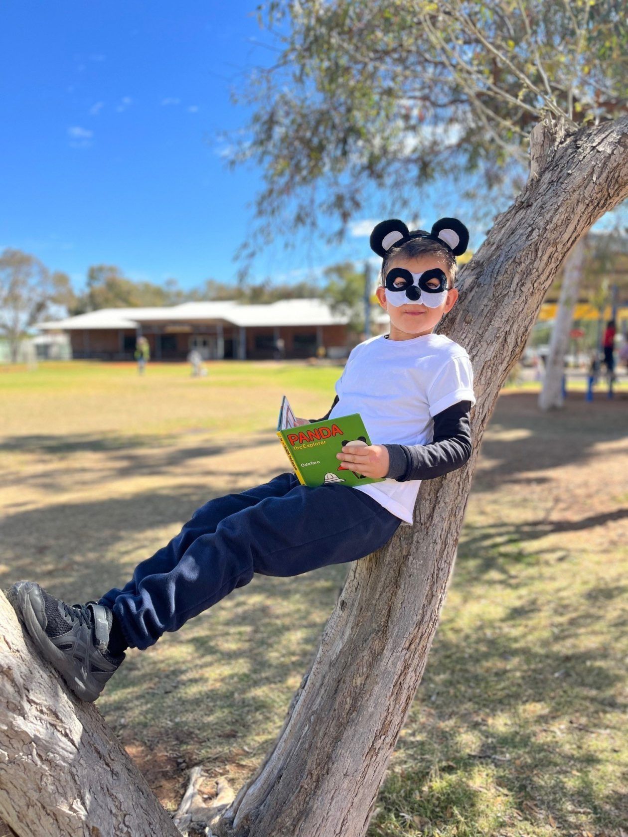 Boy in panda costume reading book in tree. Blue sky, grassy field, building in background.