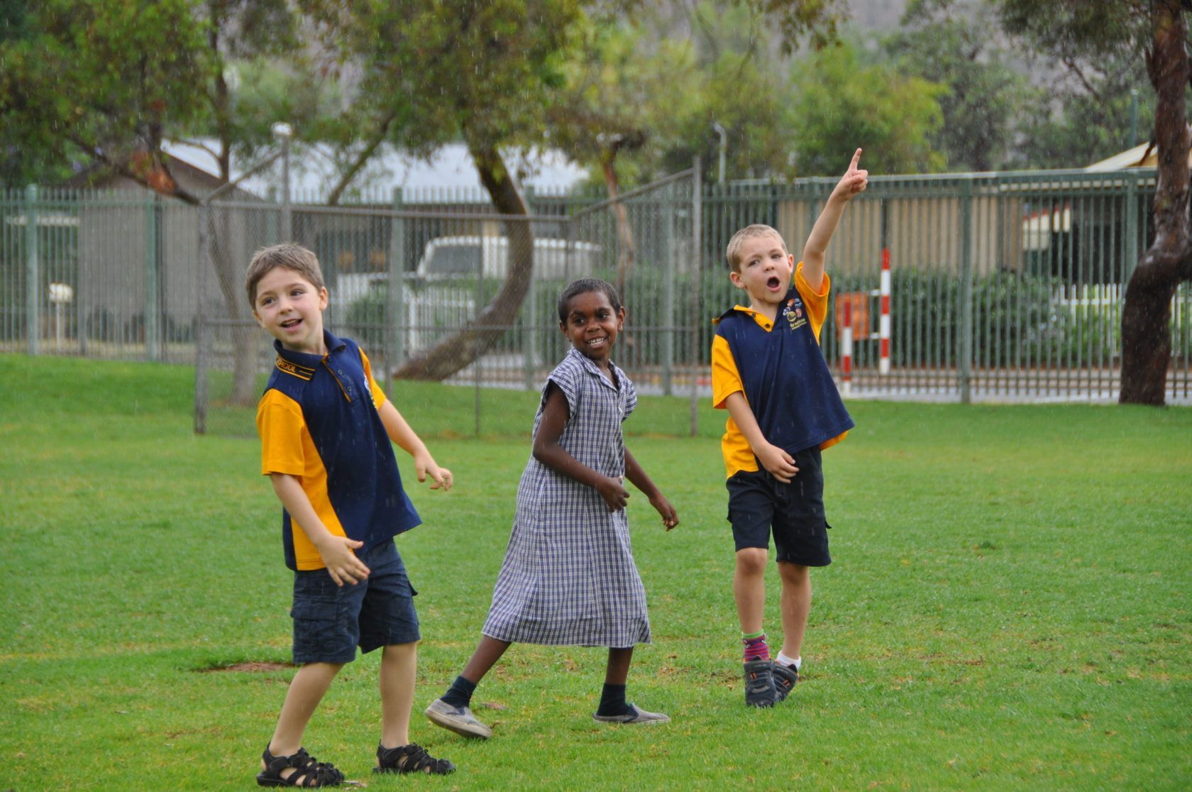 Three children playing on a grassy field; two boys in blue and yellow uniforms, one pointing, one girl in a plaid dress.